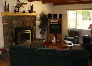 A cozy living room with a stone fireplace, a television on a wooden stand, and a large window. - 865 Michael Dr. (South Lake Tahoe)