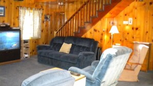 A cozy living room with a dark blue sofa, a light blue armchair, and a television on a wooden stand. A staircase with a wooden railing leads to an upper level. - 3663 Forest Ave (South Lake Tahoe)