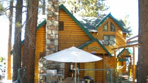 A rustic log cabin with a stone chimney, a dining area with outdoor furniture, and a large umbrella providing shade. - 3663 Forest Ave (South Lake Tahoe)