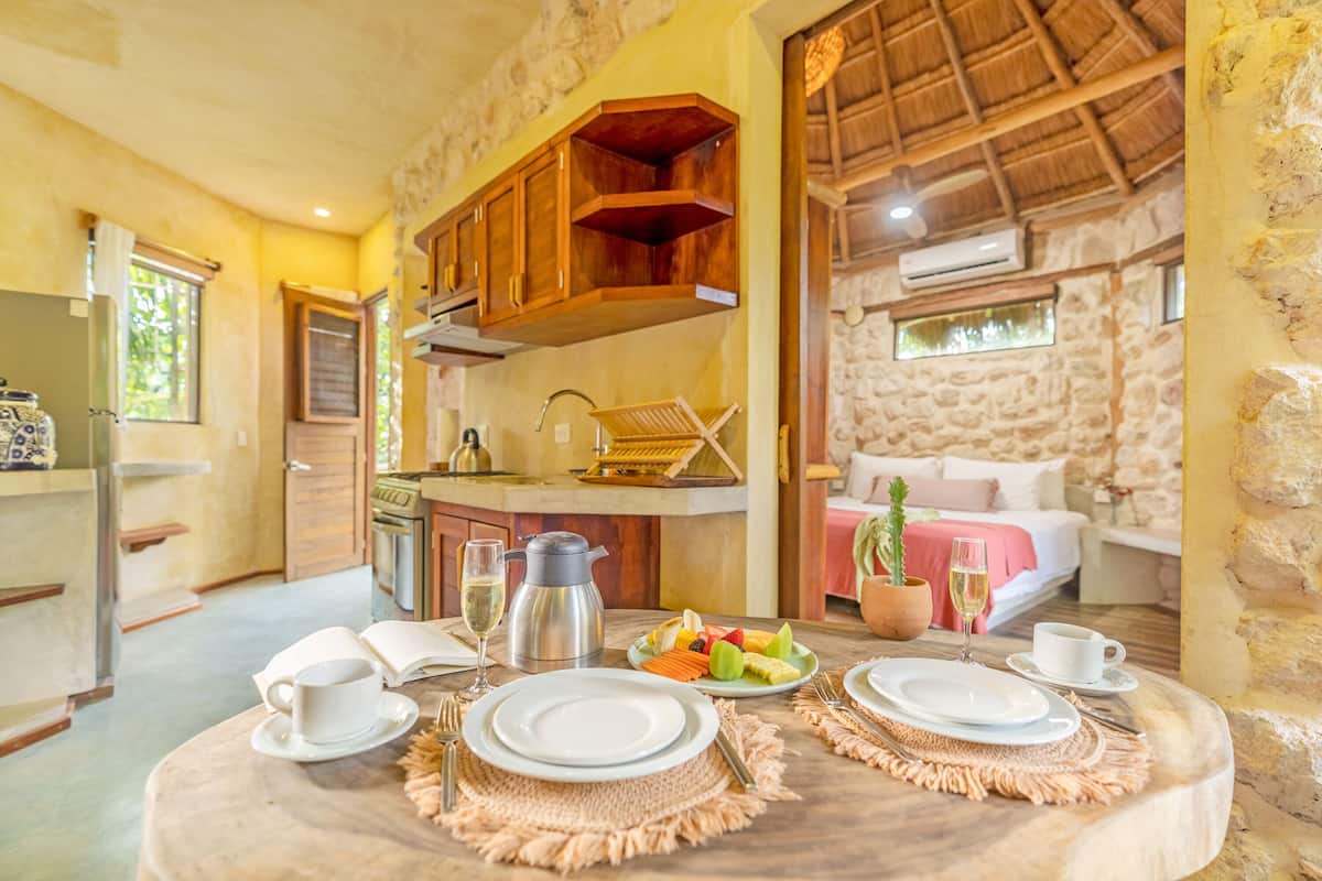 Una habitación acogedora con pared de piedra, techo de madera y cama con manta rosa. Hay una mesa de comedor con platos, tazas y un frutero.