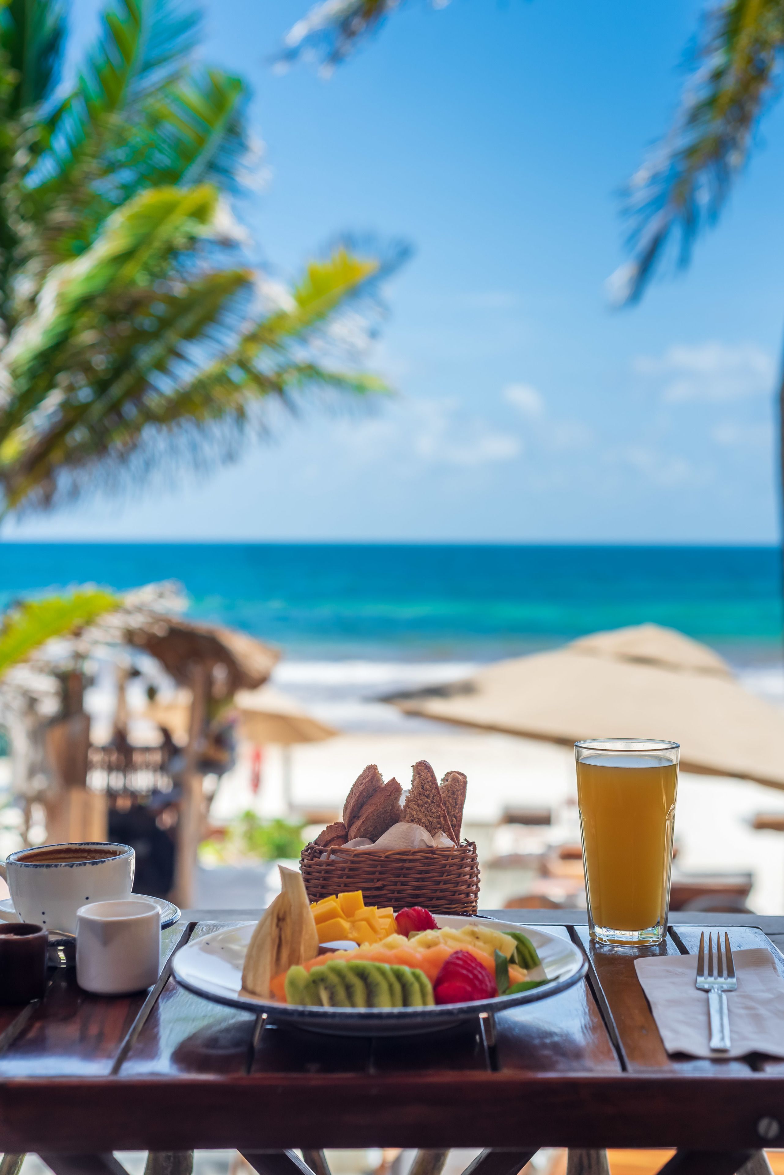 Una tavola per la colazione con un piatto di frutta, un bicchiere di succo d'arancia e una tazza di caffè, con vista su una spiaggia e l'oceano, con palme.