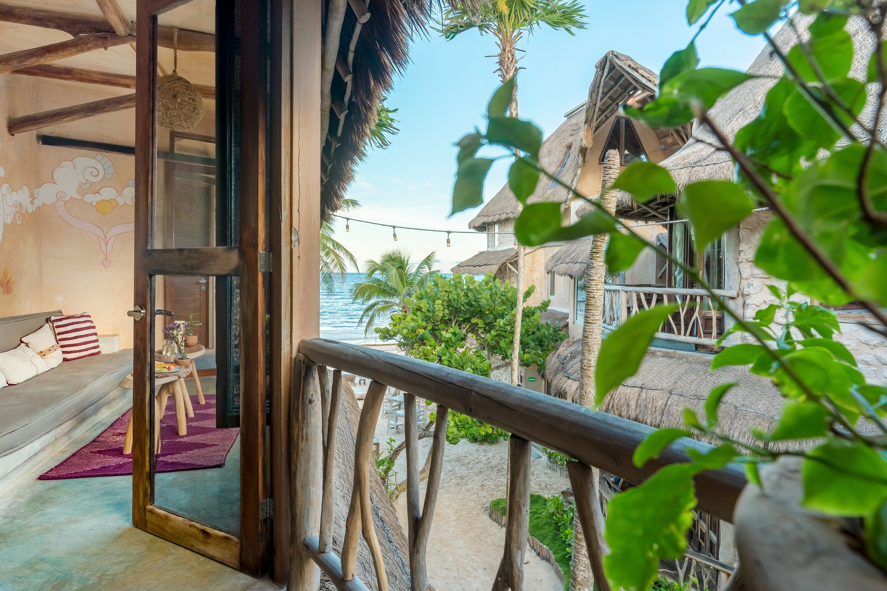 A room with a view of a beach, a wooden balcony, and a thatched-roof structure.