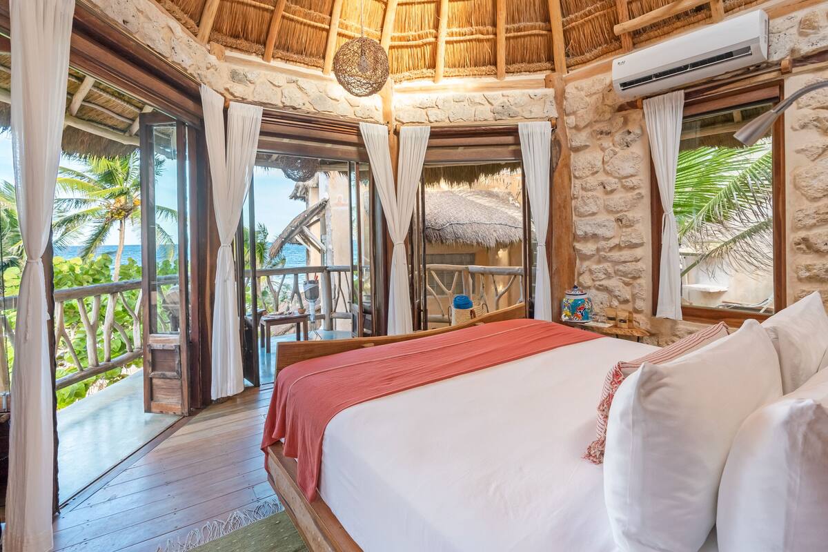 Un dormitorio con una cama grande, piso de madera, paredes de piedra y vistas a palmeras y una estructura de techo de paja.