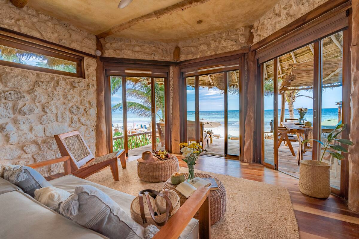 Habitación con pared de piedra, muebles de madera, vista a la playa y un comedor con mesa y sillas.