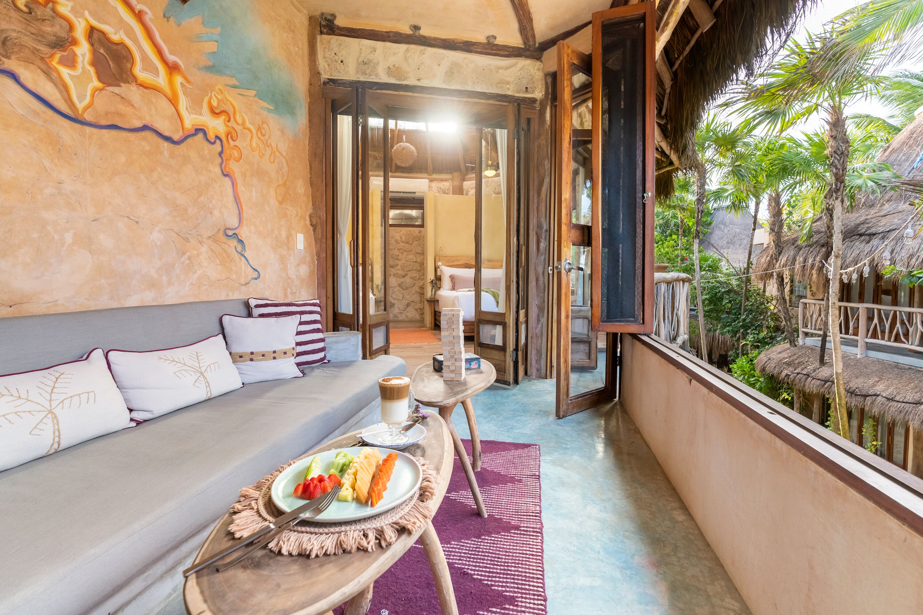A cozy room with a sofa, a dining table with a plate of fruit, and a view of a tropical garden.