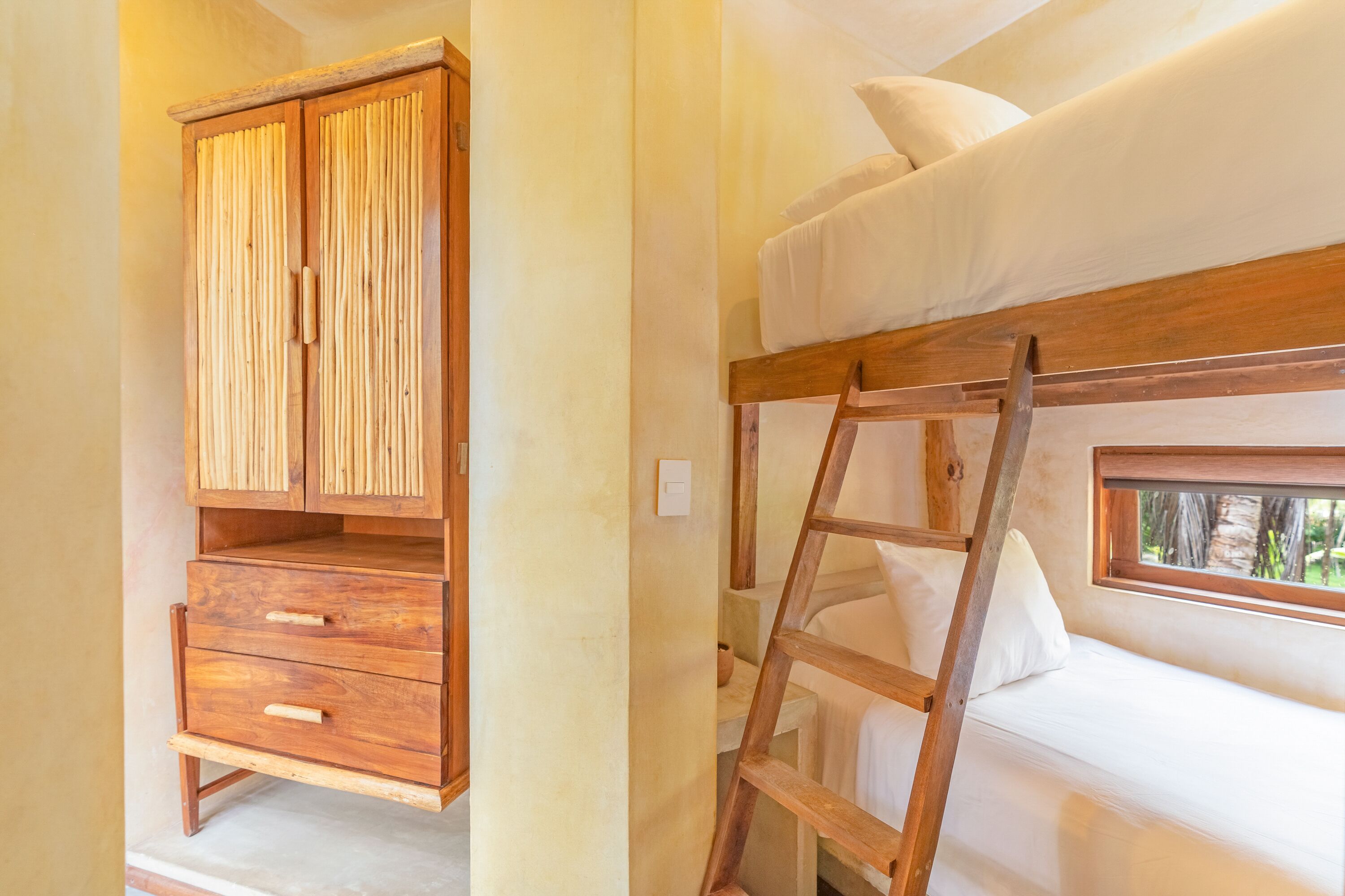 A bunk bed with a ladder, a wooden wardrobe, and a window with a view of greenery.