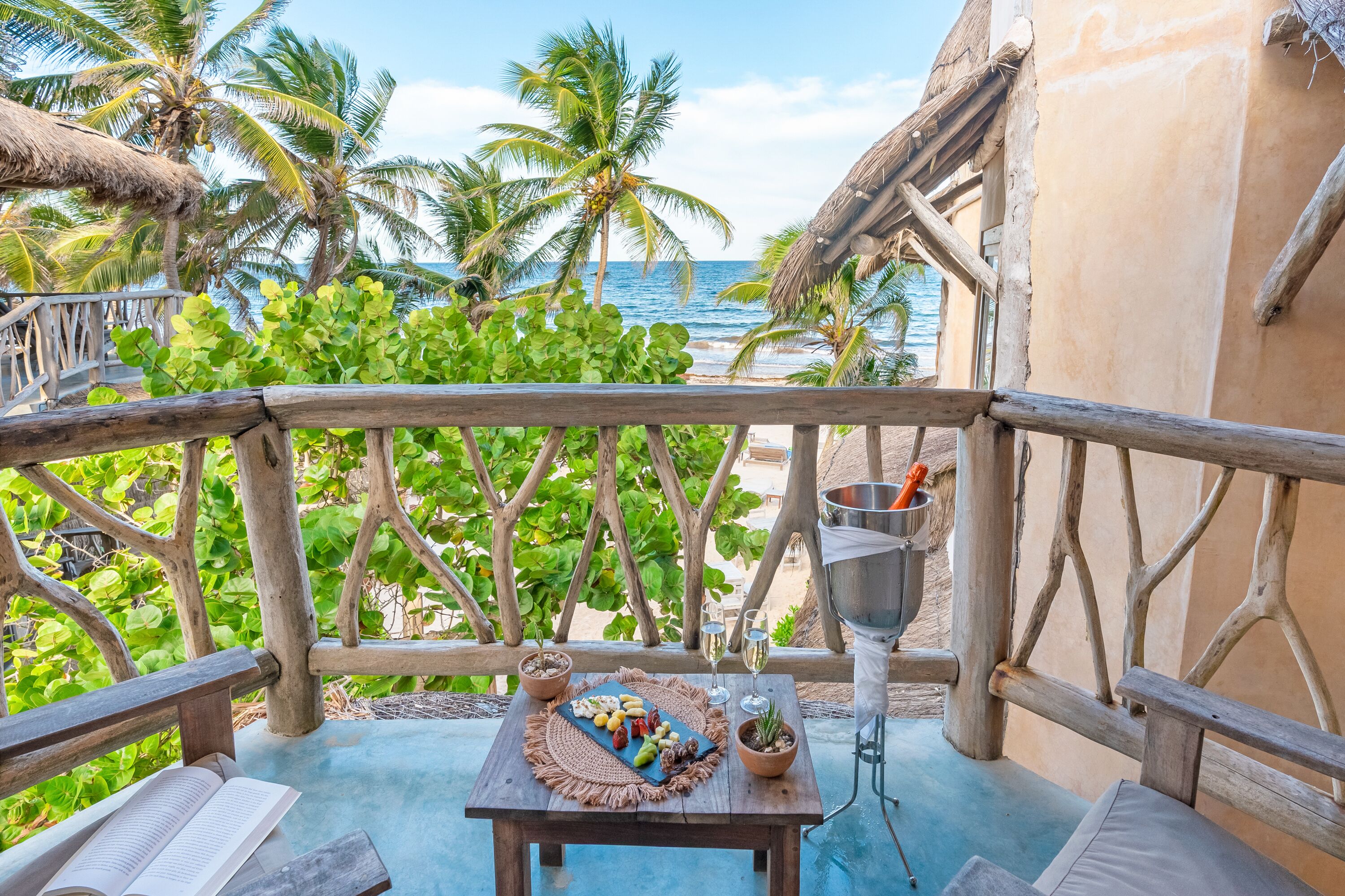 A wooden deck with a table set for two, overlooking a beach with palm trees and the ocean.