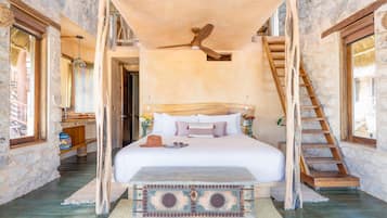 A bedroom with a large bed, a wooden bench, a ceiling fan, and a stone wall.