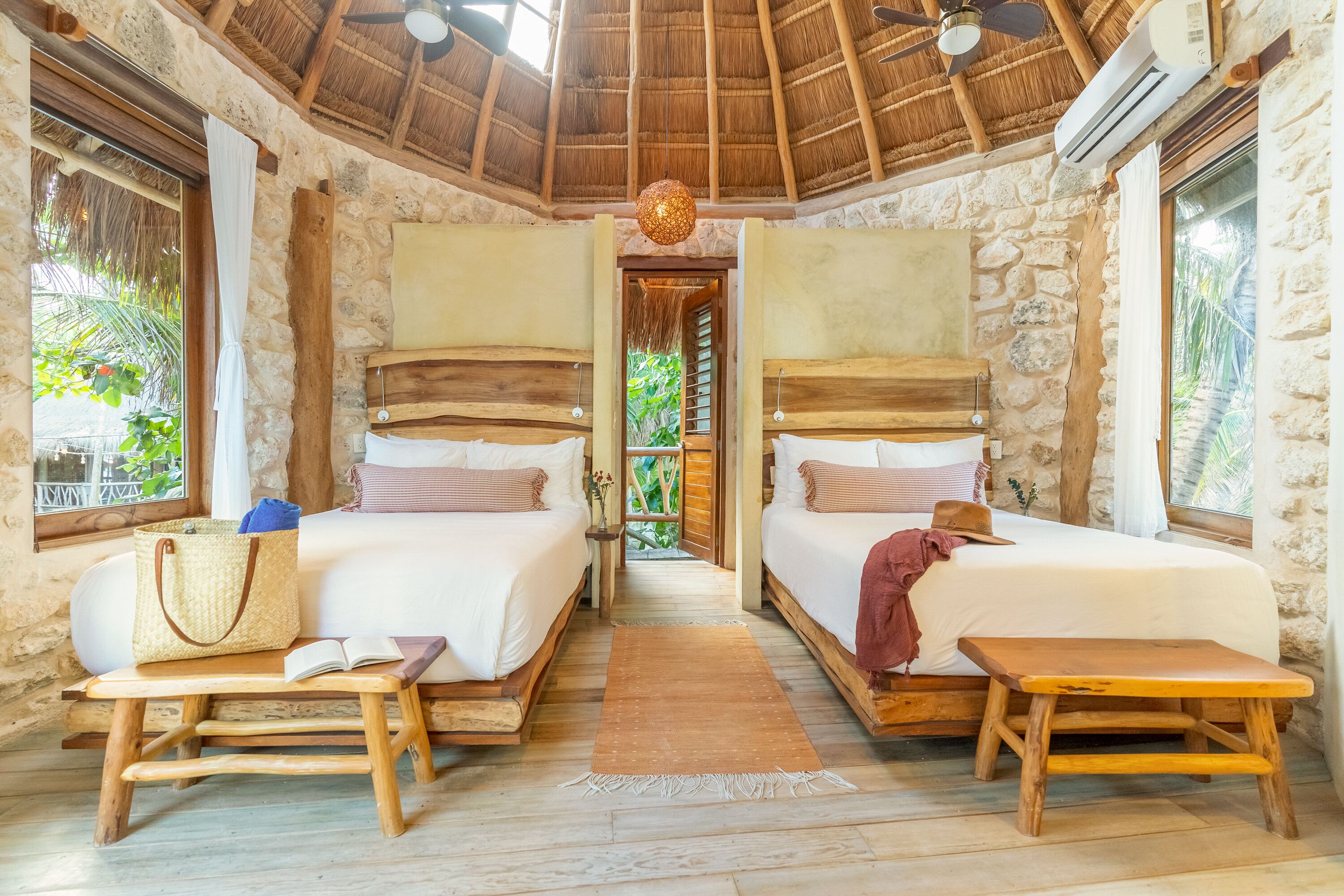 A room with two beds, a wooden bench, a straw bag, and a view of greenery.