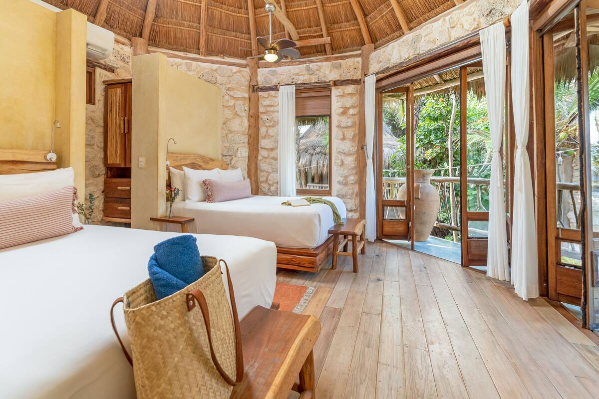 Una habitación con dos camas, un banco de madera, un bolso tejido y una pared de piedra.
