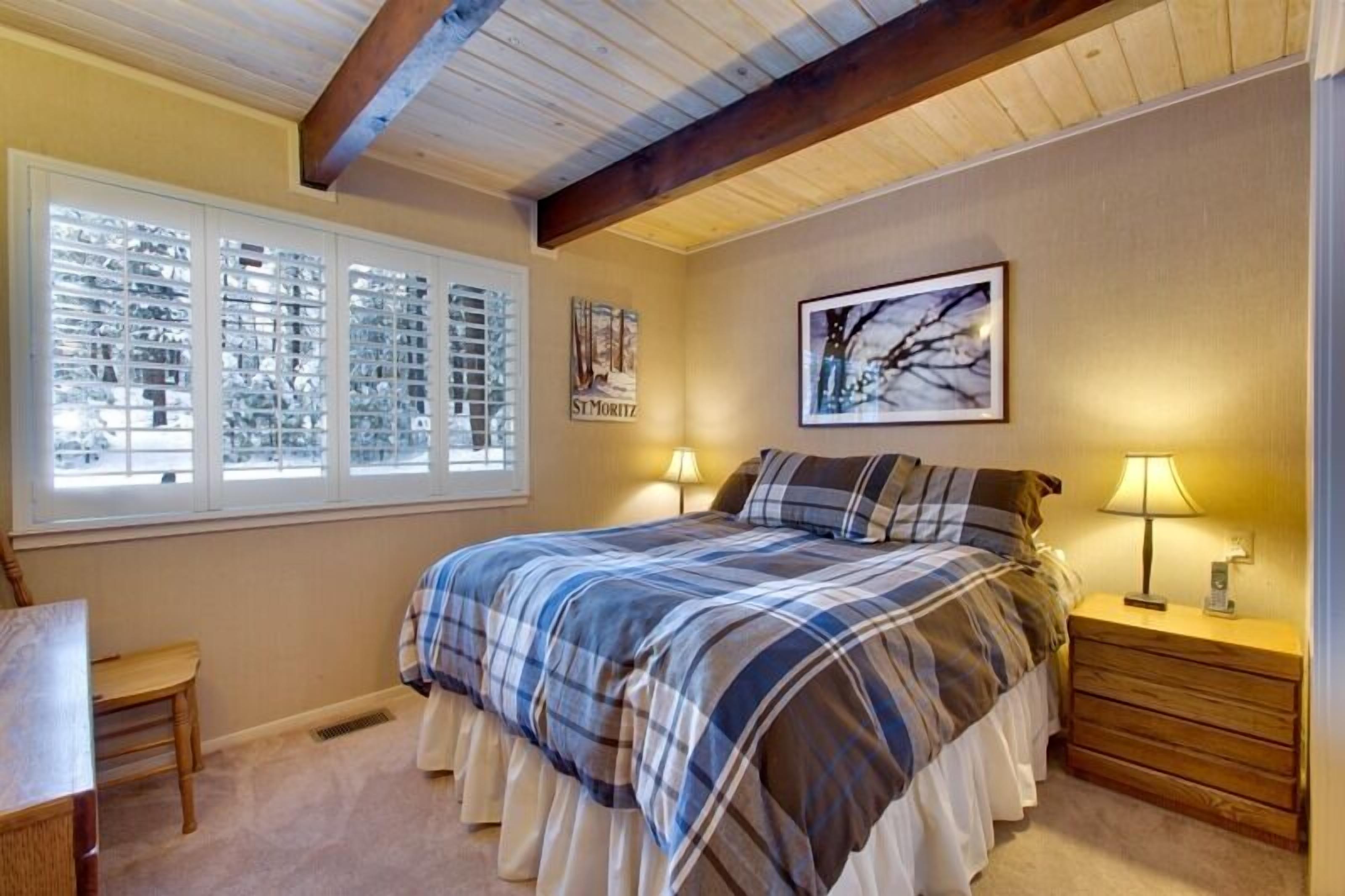 A bedroom with a large bed, two bedside lamps, a window with blinds, a framed picture on the wall, and a small wooden nightstand.