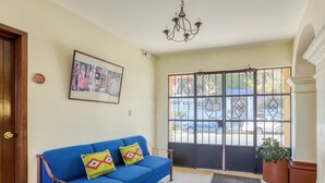 Un sofá azul con cojines coloridos, una araña, una foto enmarcada en la pared y una puerta con reja de metal.