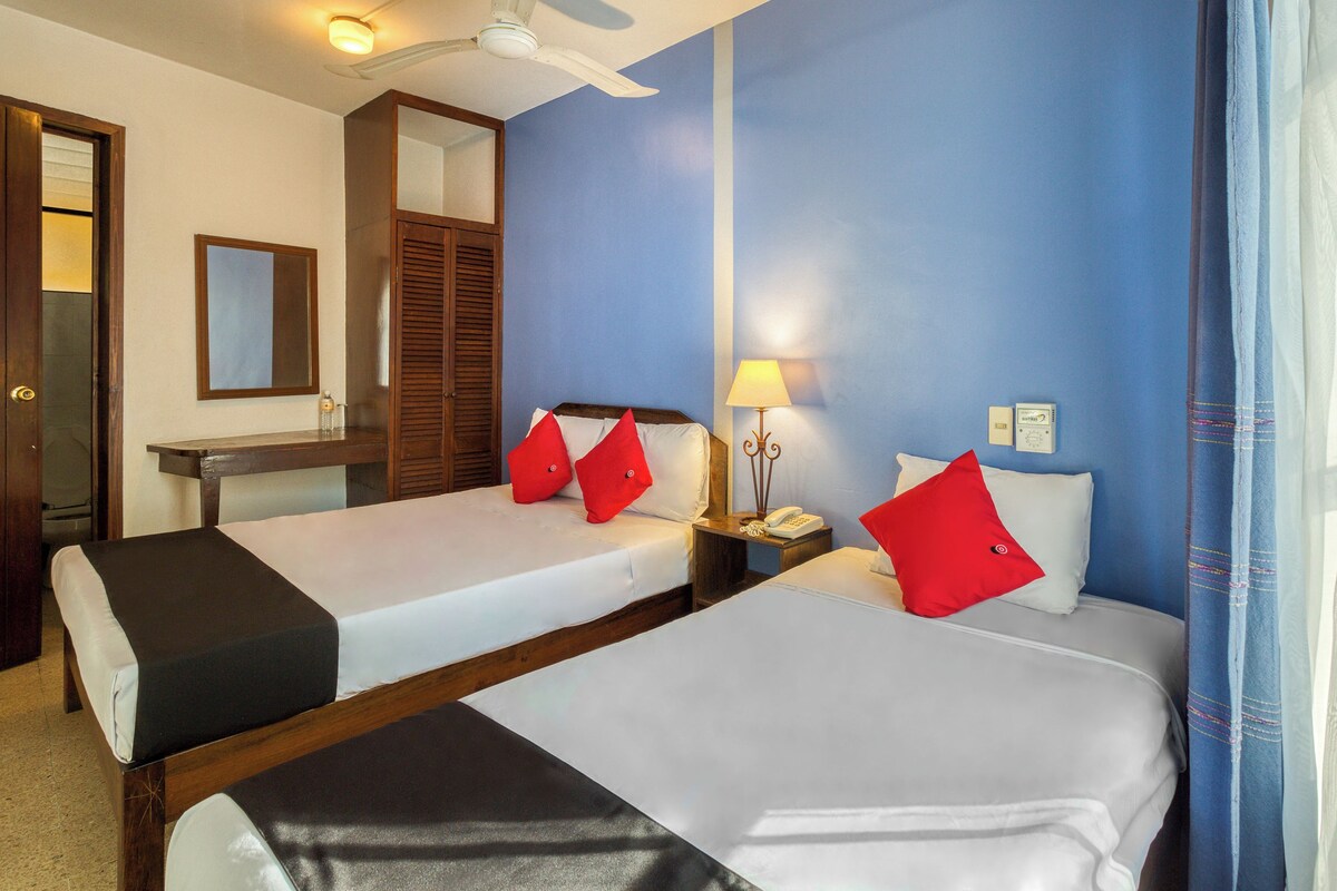 A hotel room with two beds, a wooden wardrobe, a mirror, a ceiling fan, a bedside table with a lamp, and a window with curtains.