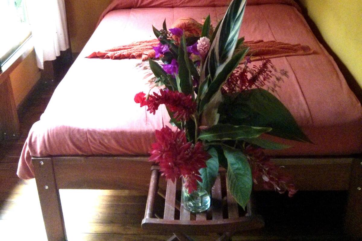 A bed with a pink bedspread, a small table with a vase of flowers, and a wall with a decorative piece.
