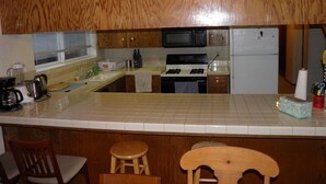A kitchen with wooden cabinets, a microwave, a stove, and a refrigerator. There are two stools and a chair. - 835 Paloma Ave. (South Lake Tahoe)