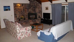 A living room with a stone fireplace, a sofa, a television, and a window with curtains. - 835 Paloma Ave. (South Lake Tahoe)