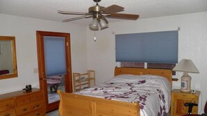 A bedroom with a wooden bed, a ceiling fan, a desk with a chair, a lamp, and a window with blinds. - 835 Paloma Ave. (South Lake Tahoe)