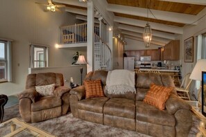 A spacious living room with a brown leather sofa set, a small table, and a lamp. A staircase leads to a loft area with a railing.