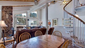 A spacious living area with a wooden dining table, comfortable seating, a stone accent wall, large windows, and a spiral staircase. - 3851 Saddle Road (South Lake Tahoe)