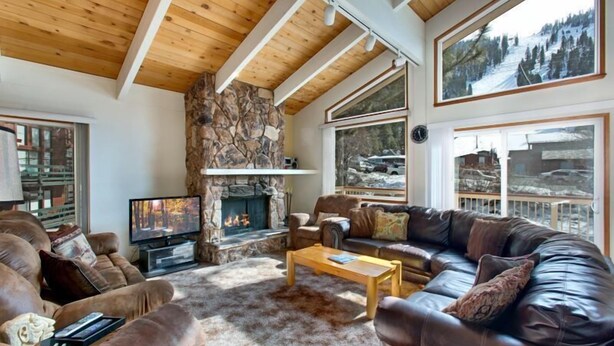A cozy living room with a stone fireplace, leather sofas, a wooden coffee table, and large windows offering scenic views. - 3851 Saddle Road (South Lake Tahoe)