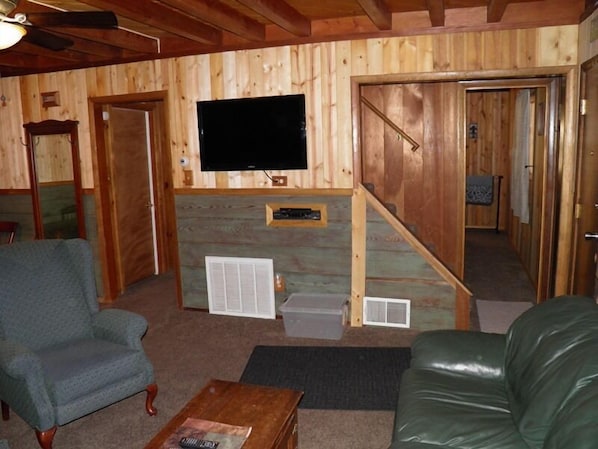A wooden interior with a television mounted on the wall, a staircase, a green leather couch, and a wooden coffee table.