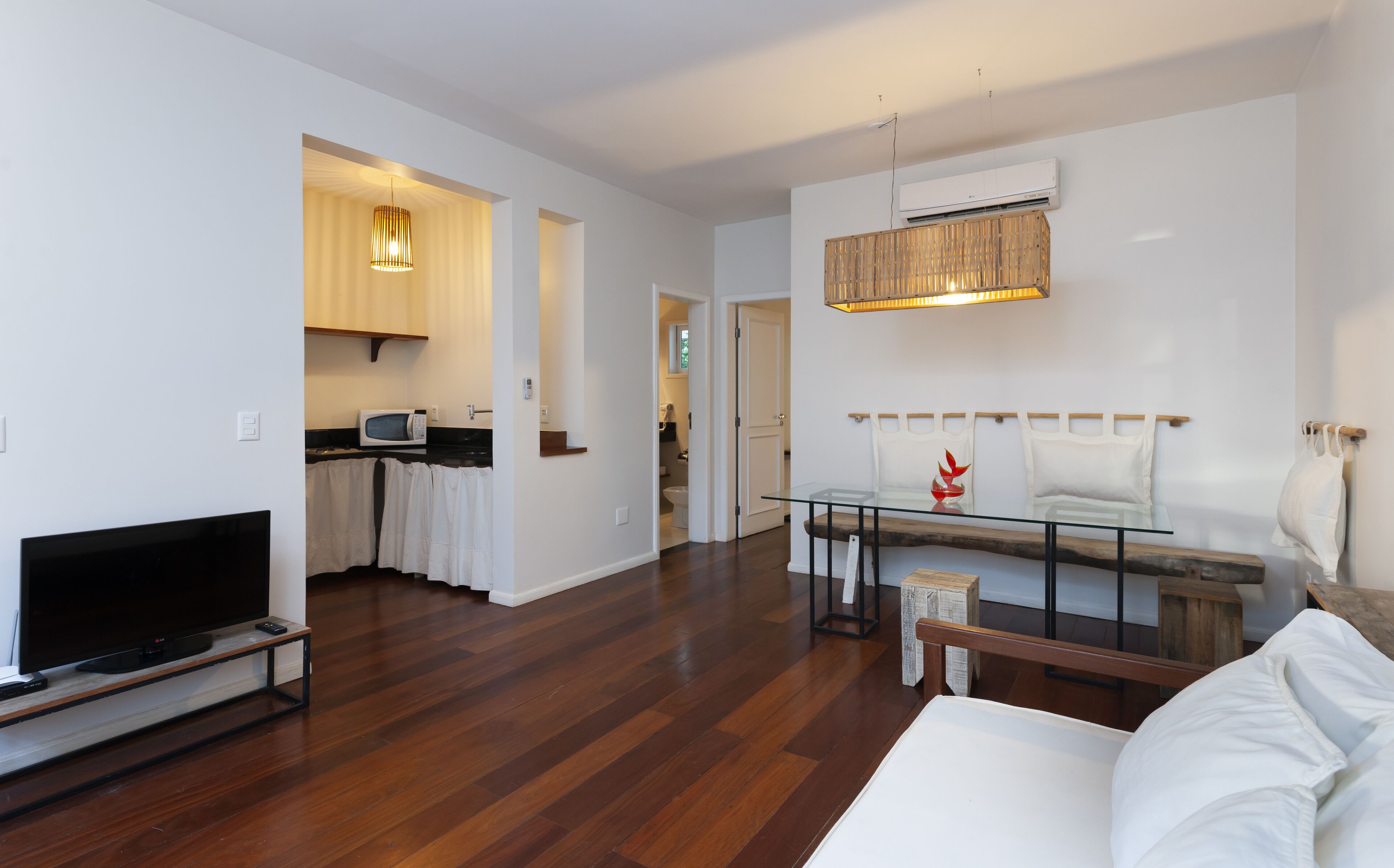 Een moderne woonkamer met een flatscreen televisie, een glazen salontafel en een bank. Er is een keukenhoek met een magnetron en een houten kast.