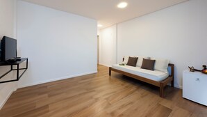 Un salon moderne avec un plancher en bois, un canapé blanc agrémenté de coussins, un petit téléviseur posé sur un meuble noir et une armoire blanche ornée d’un logo.