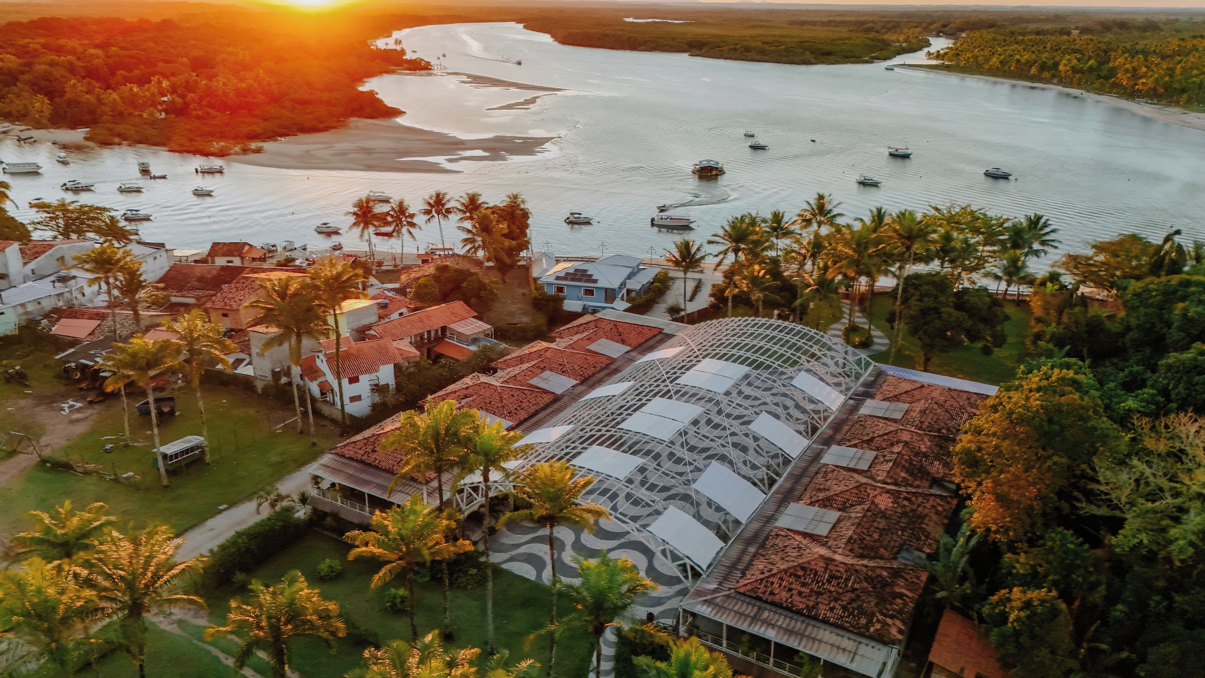 Uma área costeira com barcos na água, um edifício com telhado vermelho e palmeiras.
