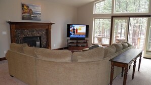 A living room with a large L-shaped sectional sofa, a fireplace, a television, and large windows with a view of trees. - 3354 Red Lake Rd (South Lake Tahoe)