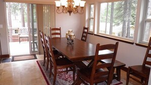 A dining room with a large wooden table, six chairs, a chandelier, and large windows with a view of trees. - 3354 Red Lake Rd (South Lake Tahoe)