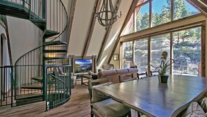 A spacious loft with a wooden floor, a spiral staircase, a dining area with a large table, and a view of the outdoors through large windows.