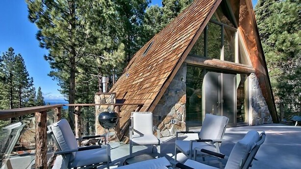 A modern A-frame cabin with a wooden exterior, large windows, and a deck with white lounge chairs.