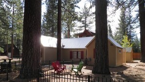 A rustic cabin with a metal roof, surrounded by tall trees. There are two red chairs and one green chair outside. - 780 Merced Ave. (South Lake Tahoe)