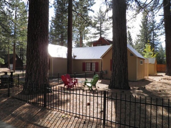 A rustic cabin with a metal roof, surrounded by tall trees. There are two red chairs and one green chair outside. - 780 Merced Ave. (South Lake Tahoe)