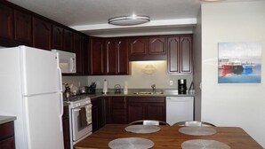 A compact kitchen with dark wooden cabinets, a white refrigerator, a stove, and a sink. There is a wooden dining table with four chairs and placemats. - 357 Ala Wai Unit 205 (South Lake Tahoe)