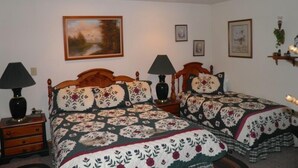 A bedroom with two wooden beds, each with a floral-patterned bedspread. There are two bedside tables with lamps, a painting on the wall, and a small shelf with decorative items.