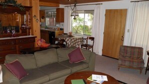 A cozy living room with a sofa, a coffee table, and a plaid armchair. There's a kitchen in the background with a dining table and chairs. - 777 Merced Ave (South Lake Tahoe)