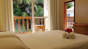 Une chambre d’hôtel avec un grand lit, une chaise en bois, une petite table et un balcon donnant sur des palmiers.