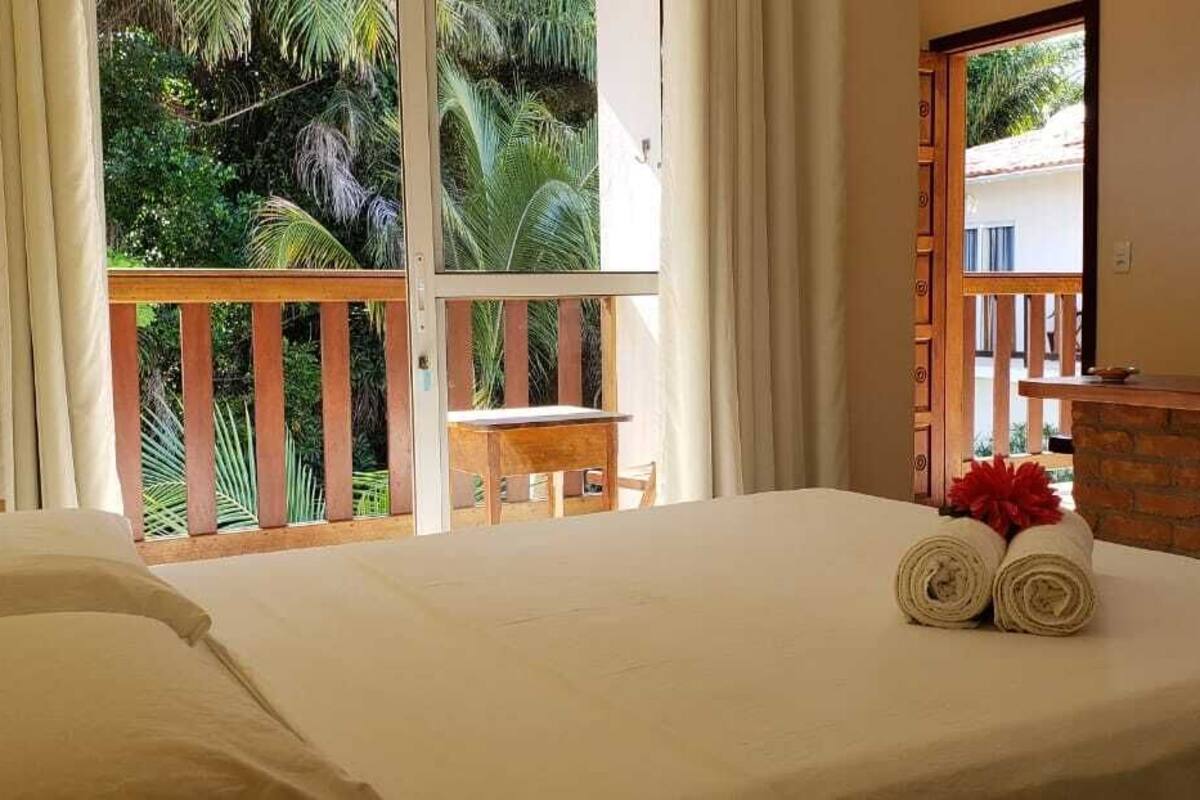 Habitación de hotel con una cama grande, una silla de madera, una mesita pequeña y un balcón con vistas a palmeras.