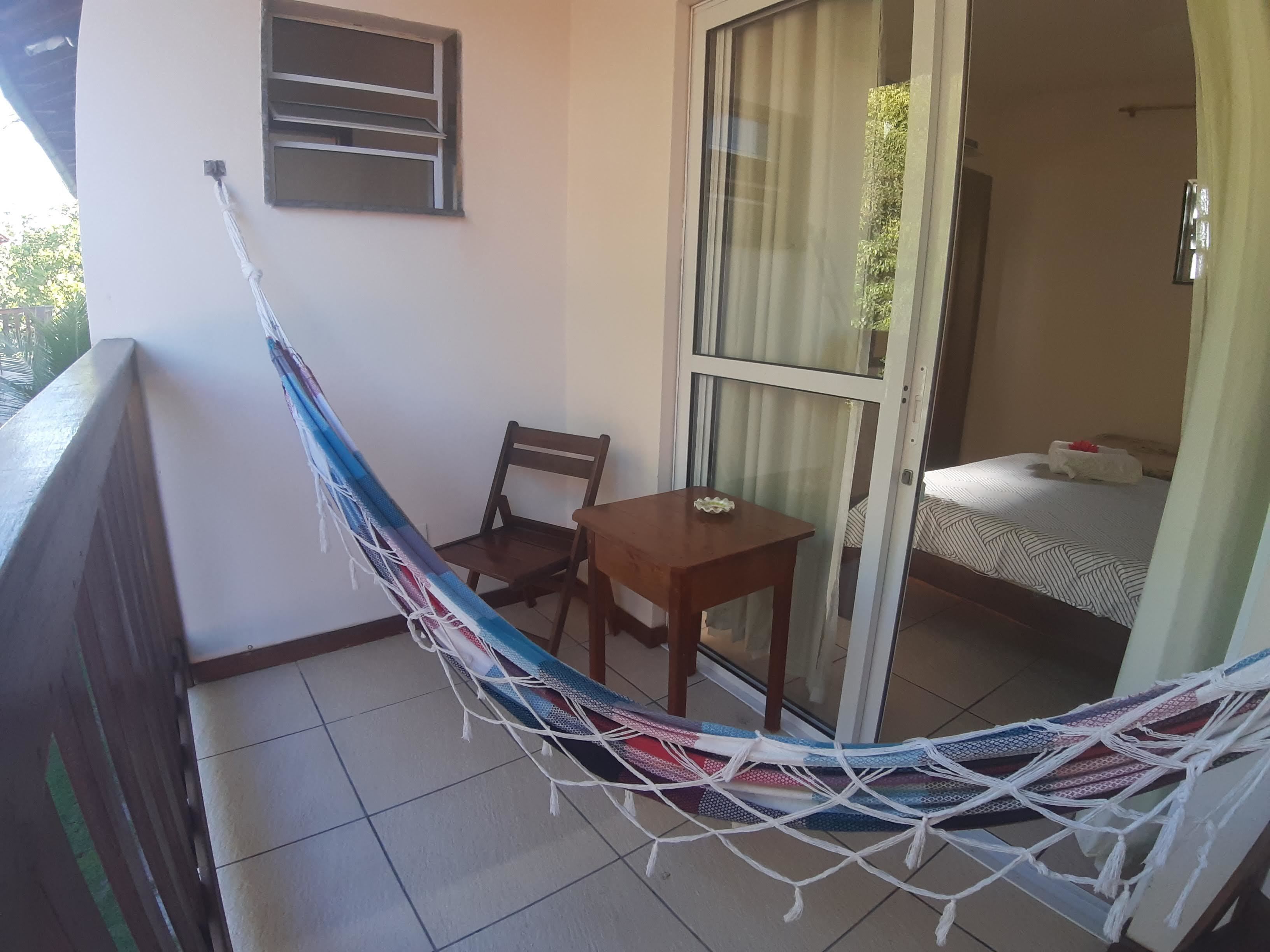 Un balcón con una hamaca, una silla de madera, una mesita pequeña y una cama con colcha a rayas.