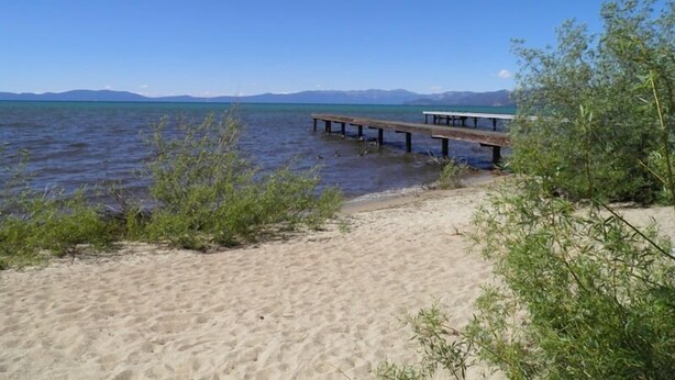 En sandstrand med en träbrygga som sträcker sig ut i stilla vatten, omgiven av grönska och berg i fjärran.