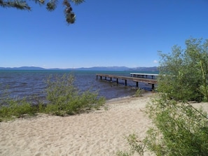 A sandy beach with a wooden pier extending into calm water, surrounded by greenery and mountains in the distance.