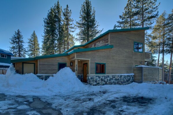 A two-story log cabin with a stone foundation, surrounded by snow and tall pine trees.