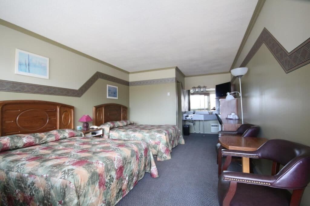 Quarto de hotel com duas camas, uma escrivaninha, uma cadeira e um banheiro ao fundo.