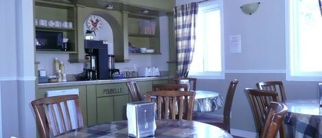 A dining area with a round table covered in a checkered tablecloth, surrounded by wooden chairs. A kitchen with green cabinets and a coffee machine is visible in the background.