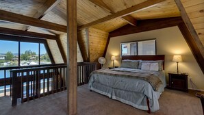 A bedroom with a large bed, two bedside lamps, a wooden ceiling, and a view of a body of water.