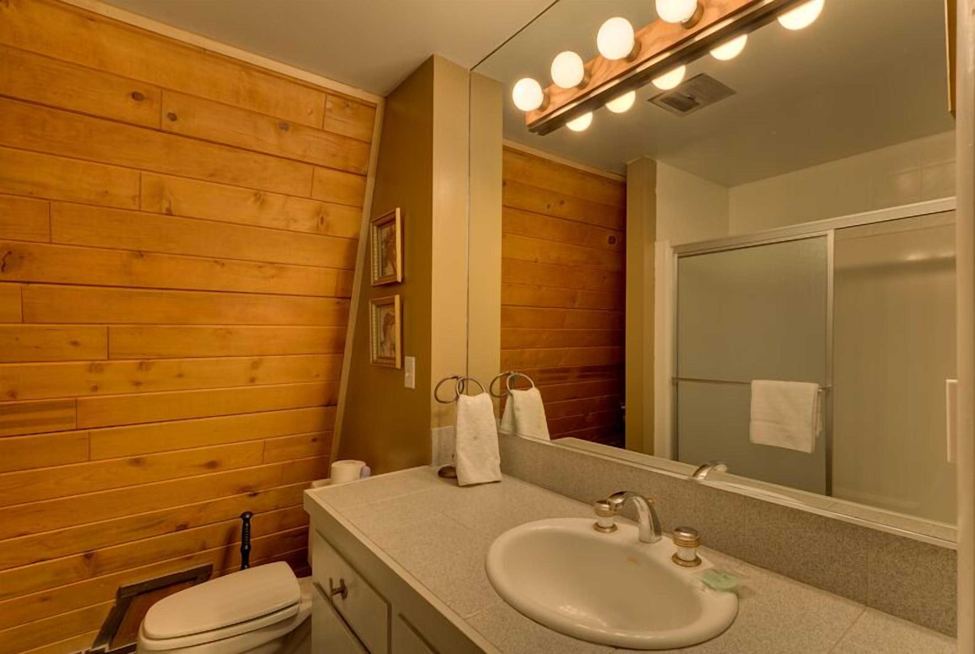 A bathroom with wooden walls, a sink, a mirror with lights, and a shower.
