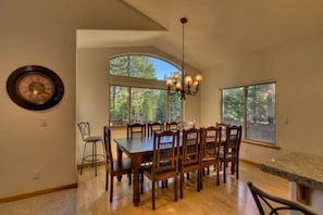 A dining area with a large wooden table and chairs, a chandelier, and large windows offering a view of trees.
