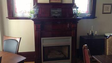 A room with a fireplace, a dining table, and chairs. There is a plaque above the fireplace with a name and date.
