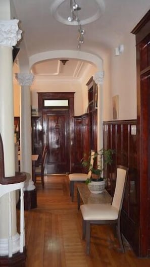 Un pasillo con piso de madera, una silla blanca, una mesa con planta en maceta y una puerta de madera al final.